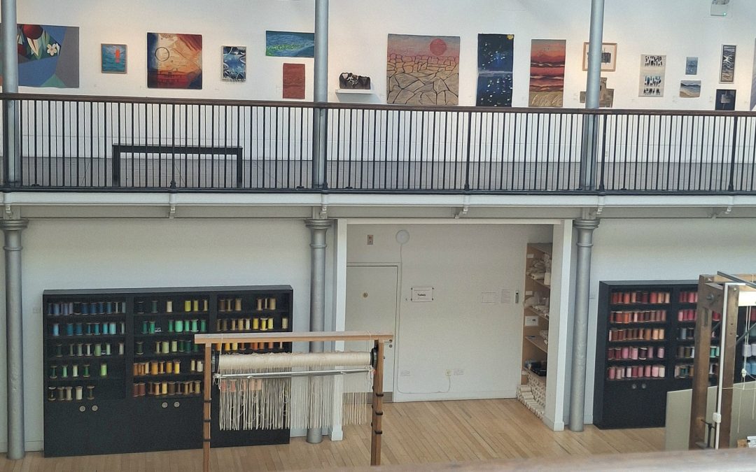 A high-angle view of the Dovecot Studios weaving floor and gallery balcony. Below, shelves are stocked with colorful spools of yarn next to a large upright loom. Above, an exhibition of contemporary woven tapestries by the British Tapestry Group is displayed along a white gallery wall.