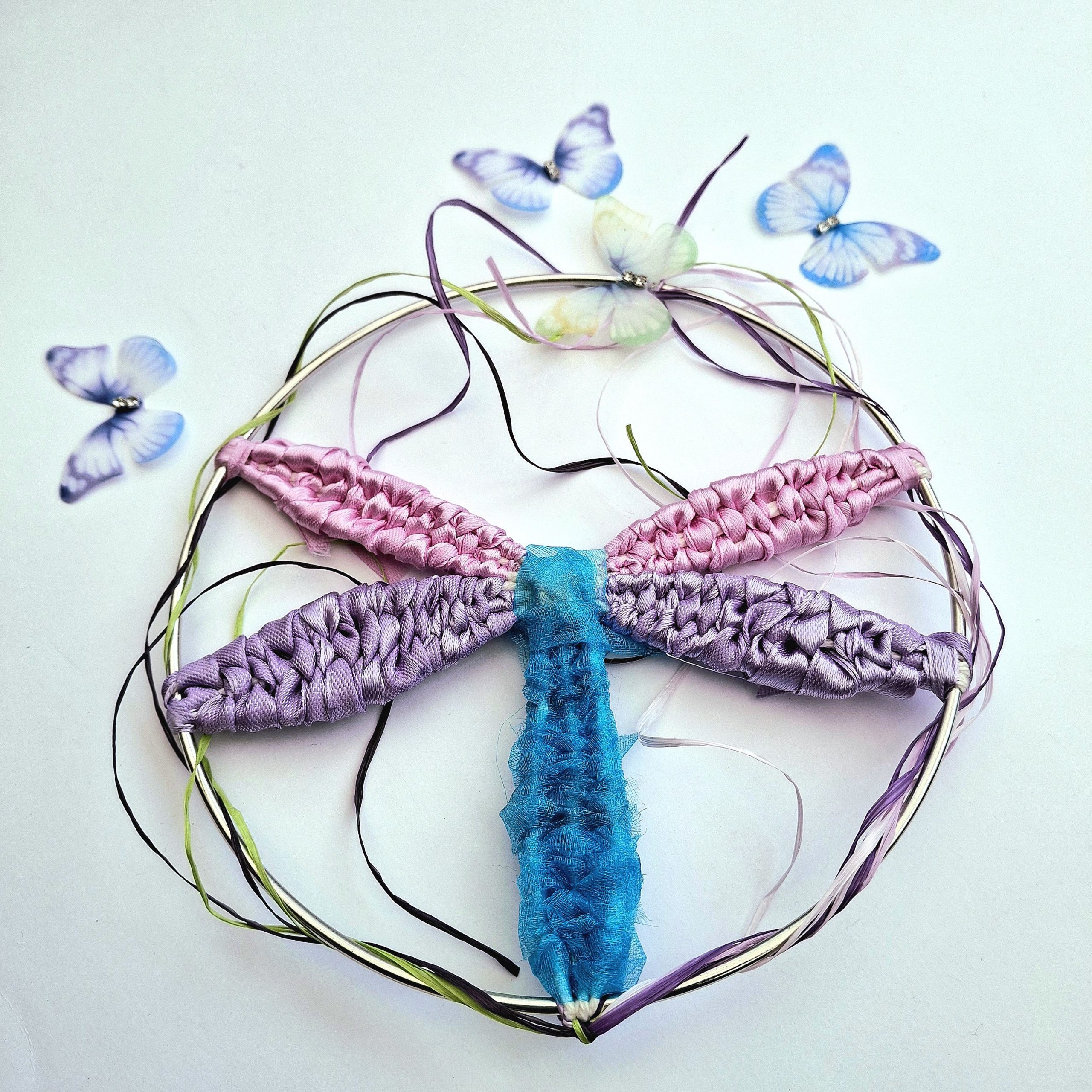 A handmade woven dragonfly on a silver metal hoop. The dragonfly features a bright blue textured body and four woven wings in shimmering pink and purple ribbons, surrounded by delicate decorative butterflies.