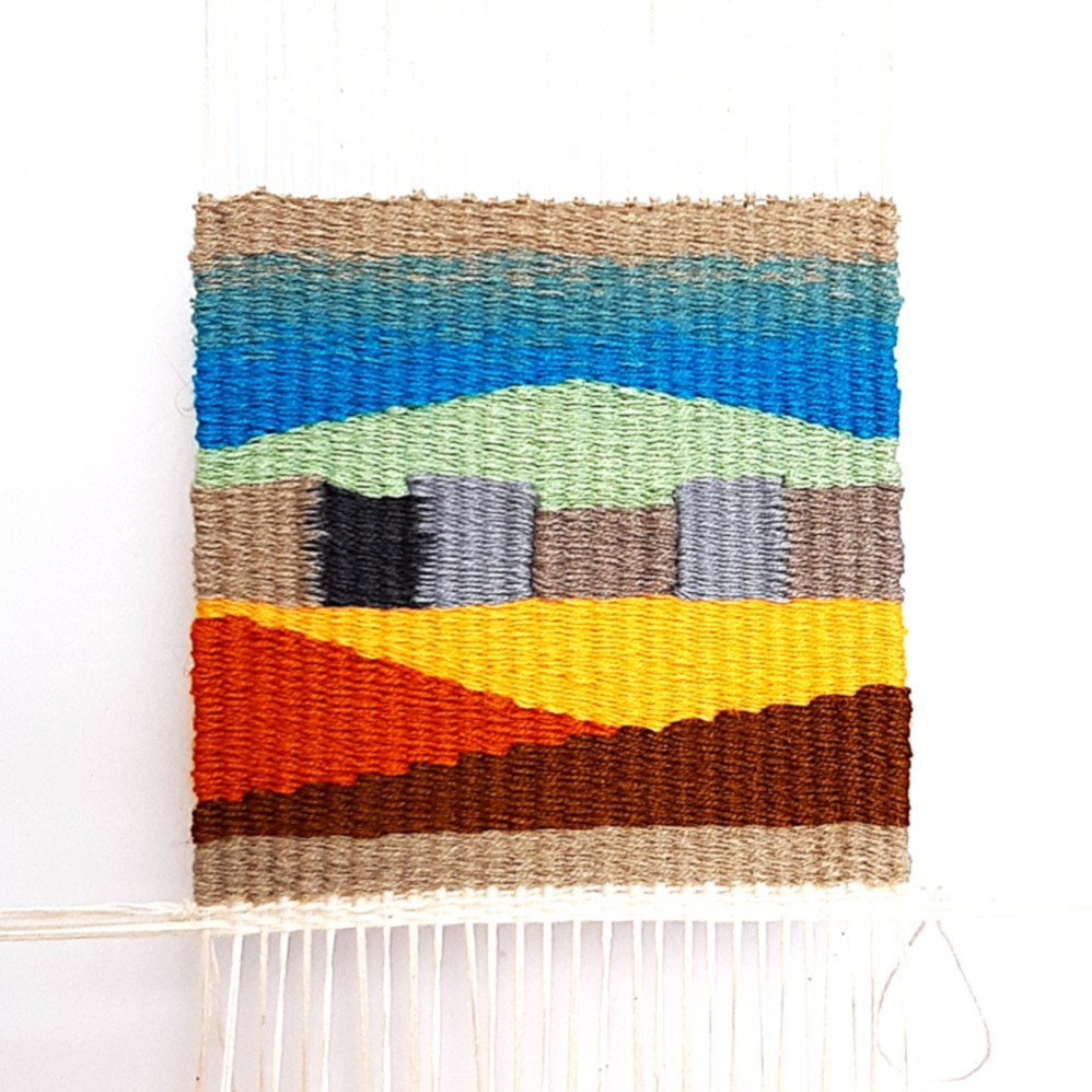 A small, hand-woven tapestry on a loom featuring a geometric landscape with a blue sky, green hills, and warm yellow and terracotta fields, demonstrating the weft-faced weaving technique.