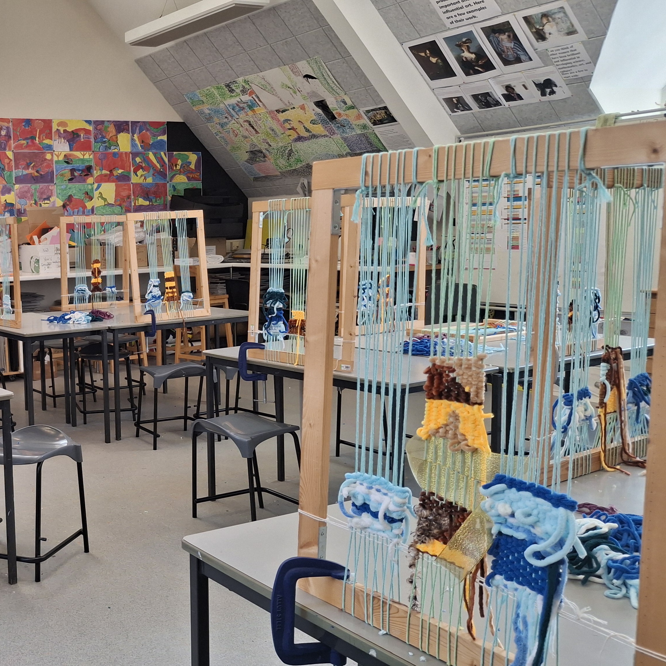 A bright art classroom set up for a textile workshop, featuring several large wooden frame looms clamped to desks, each prepared with light blue warp threads and colourful yarn for students.